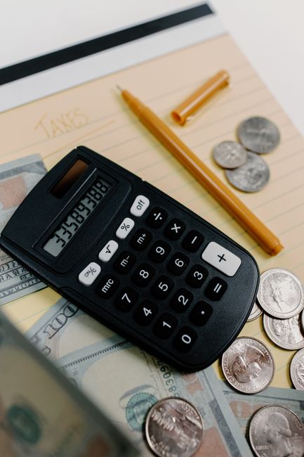 A photograph of a home packing and moving setup on a cardboard surface. Visible items include a calculator with white and pink buttons, a roll of black tape, a set of multi-coloured file folders or no