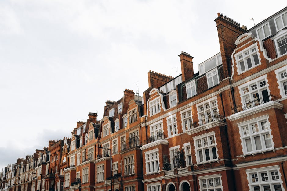 Photograph capturing the upper floors of a row of historic red brick townhouses on Sloane Street, Knightsbridge, with multiple white-framed sash windows, some featuring small balconies with black wrought-iron railings, and chimney stacks visible on the roofline. The buildings are closely aligned, exhibiting Victorian architectural details, against an overcast sky. In the foreground, the lower part of the buildings shows a tiled sidewalk and street-level entrances, with no vehicles or people present. This scene reflects the typical residential exterior in the Knightsbridge area, relevant to a house removal context, where furniture, boxes, and packing materials from apartment interiors would be transported during a home relocation or furniture transport process, often involving loading onto a removals van managed by companies such as Removals Knights Bridge.
