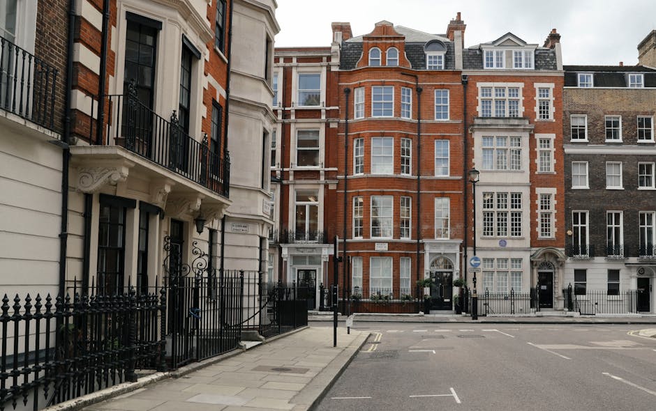 A straight-on street view of tall, multi-storey residential buildings in Knightsbridge, London, with a mix of brick and white facades. The pavement features black wrought iron fences and small gate entrances leading to the buildings’ ground-floor apartments. The street is empty, with a few parking bays and road markings visible, and the sky is overcast. In the foreground, a logistics process is inferred from the context, as the image relates to home relocation services; this includes the possible presence of a removals van parked nearby, though it is not directly visible, and the setting suggests preparation for furniture transport or packing and moving activities associated with house removals, with a focus on the exterior environment suitable for the initial stages of a residential move, as handled by Removals Knights Bridge.