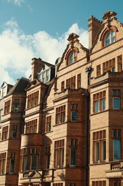 Photograph capturing the upper floors of a row of historic red brick townhouses on Sloane Street, Knightsbridge, with multiple white-framed sash windows, some featuring small balconies with black wrought-iron railings, and chimney stacks visible on the roofline. The buildings are closely aligned, exhibiting Victorian architectural details, against an overcast sky. In the foreground, the lower part of the buildings shows a tiled sidewalk and street-level entrances, with no vehicles or people present. This scene reflects the typical residential exterior in the Knightsbridge area, relevant to a house removal context, where furniture, boxes, and packing materials from apartment interiors would be transported during a home relocation or furniture transport process, often involving loading onto a removals van managed by companies such as Removals Knights Bridge.